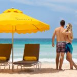 Couple walking arm-in-arm along Rockley Beach with yellow umbrellas and turquoise water